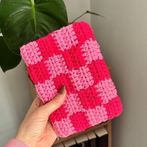Puede incluir: Un artículo cuadrado de ganchillo hecho a mano con un patrón de tablero de ajedrez en rosa brillante y rosa claro. El artículo se sostiene en una mano, con una estantería y una planta verde en el fondo. El artículo parece ser una pequeña bolsa o estuche.