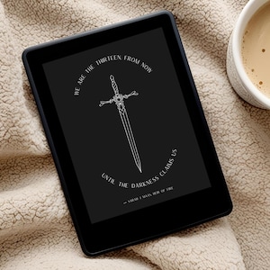 May include: A black and white e-reader with a quote on the screen that reads "We are the thirteen from now until the darkness claims us - Sarah J. Maas, House of Earth and Blood". The e-reader is on a beige fuzzy blanket.