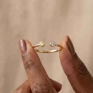 Puede incluir: Un anillo de oro con un diseño abierto, con una gema amarilla en un extremo y una gema transparente en el otro. El anillo se sostiene entre dos dedos sobre un fondo neutro.