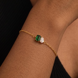 Puede incluir: Una pulsera de cadena en tono dorado con dos gemas adyacentes: una piedra rectangular de color verde esmeralda y una piedra transparente en forma de pera. La delicada cadena de metal se conecta a cada lado del engaste de las piedras.