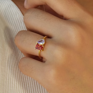 Puede incluir: Un anillo de oro con dos piedras preciosas. La piedra más grande es cuadrada y roja. La piedra más pequeña es de forma de lágrima y púrpura.