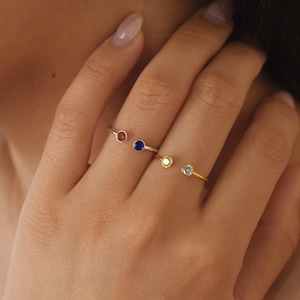 May include: Three gold rings with different coloured gemstones. The rings are open and adjustable. The gemstones are red, blue, and clear.