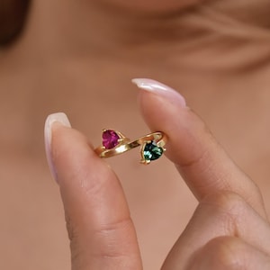 Könnte beinhalten: Ein goldener Ring mit einem rosafarbenen und grünen herzförmigen Stein. Der Ring ist so konzipiert, dass er am Mittelfinger getragen wird.