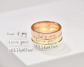 Puede incluir: Tres anillos apilados con mensajes escritos a mano. El anillo superior es de oro rosa, el anillo del medio es de oro y el anillo inferior es de plata. Los mensajes en los anillos son "love of my", "I love you" y "GREIGHTON".