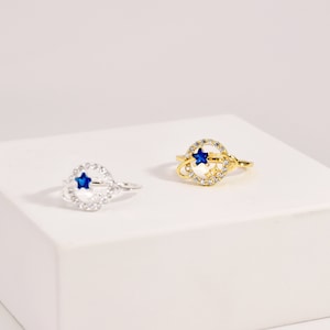 Puede incluir: Dos anillos con una estrella azul y cristales blancos en un diseño circular. El anillo de la izquierda es de plata y el anillo de la derecha es de oro.