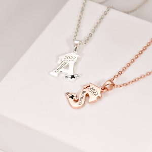 May include: Two silver necklaces with graduation cap pendants. The pendant on the left is a silver letter A with the name "Charlie" and the year "2022" engraved on it. The pendant on the right is a rose gold letter S with the name "Romina" and the year "2022" engraved on it.