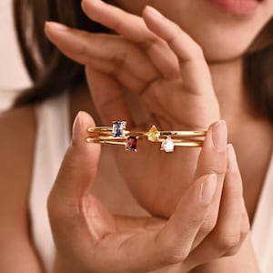 Peut inclure: Bracelets dorés, chacun orné d'une pierre précieuse différente. Les pierres précieuses comprennent une pierre bleue rectangulaire, une pierre rouge rectangulaire, une pierre jaune en forme de goutte et une pierre blanche en forme de goutte. Les bracelets sont tenus dans une main.