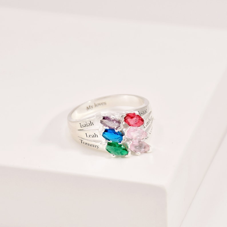 May include: A silver ring with multiple colored gemstones. The ring has the words "My loves" engraved on the band. The names "Isaiah", "Leah", "Tommy", and "Isaac" are also engraved on the ring.