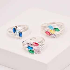 May include: Three silver rings with colorful gemstone clusters. Each ring features a different arrangement of blue, green, yellow, pink, and purple stones. Names are engraved on the bands, adding a personalized touch to these pieces of jewelry.