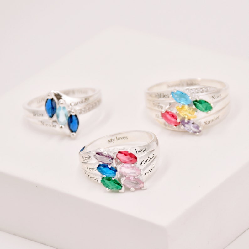 May include: Three silver rings with colorful gemstone clusters. Each ring features a different arrangement of blue, green, yellow, pink, and purple stones. Names are engraved on the bands, adding a personalized touch to these pieces of jewelry.