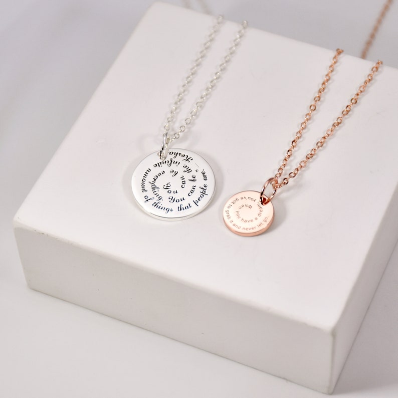 Puede incluir: Dos collares con colgantes, uno plateado y otro oro rosa, expuestos sobre una superficie blanca. El collar plateado tiene un colgante grande y redondo con texto grabado. El collar oro rosa tiene un colgante redondo m&aacute;s peque&ntilde;o con texto grabado.