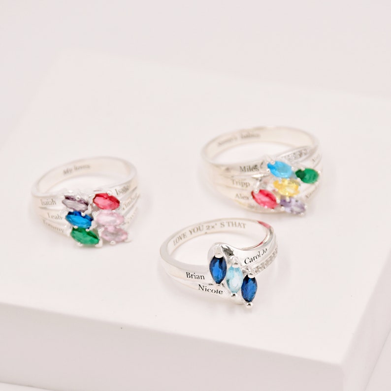 May include: Three silver rings with colorful marquise-cut gemstones. Each ring features a cluster of gems in various colors, including blue, green, pink, and yellow. The rings have engraved names and phrases.