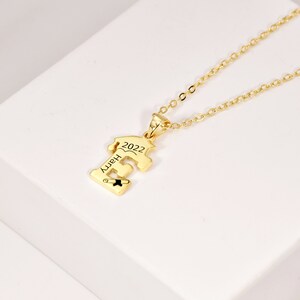 May include: Gold necklace with a graduation cap pendant. The pendant is shaped like the letter 'E' and has the year '2022' and the word 'Happy' engraved on it.
