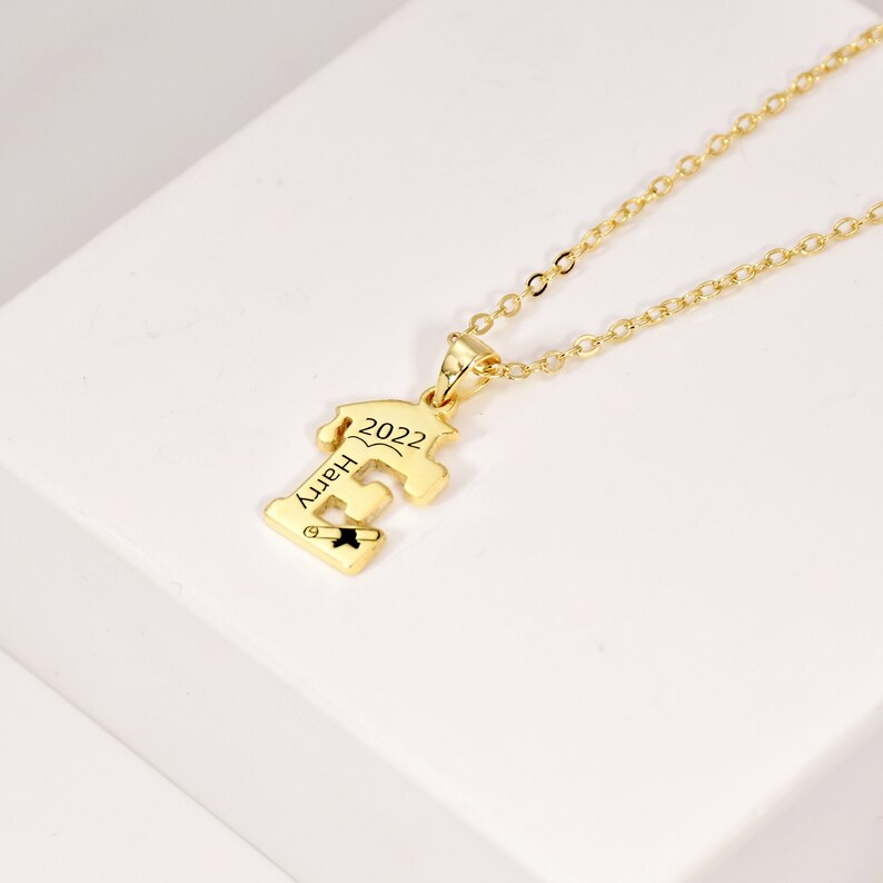 May include: Gold necklace with a graduation cap pendant. The pendant is shaped like the letter 'E' and has the year '2022' and the word 'Happy' engraved on it.
