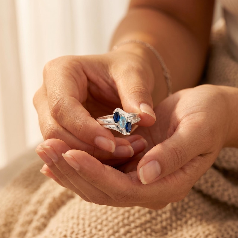 May include: A silver ring with two oval-shaped blue gemstones and smaller clear stones. The ring has the names "Brian", "Nicole", and "Carol" engraved on it. The ring is held in a person's hands.
