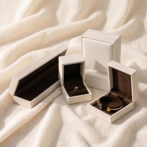 May include: A collection of white jewellery boxes with brown velvet linings. One box contains a ring, and another holds a gold bracelet with blue and yellow gemstones. The boxes are arranged on cream-coloured fabric.