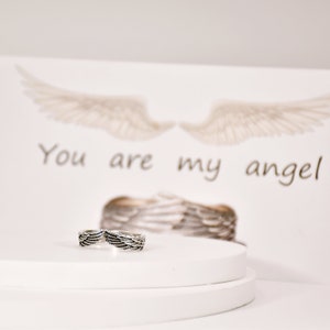 May include: A silver ring with a design of two angel wings. The ring is on a white platform with a card behind it that says "You are my angel".