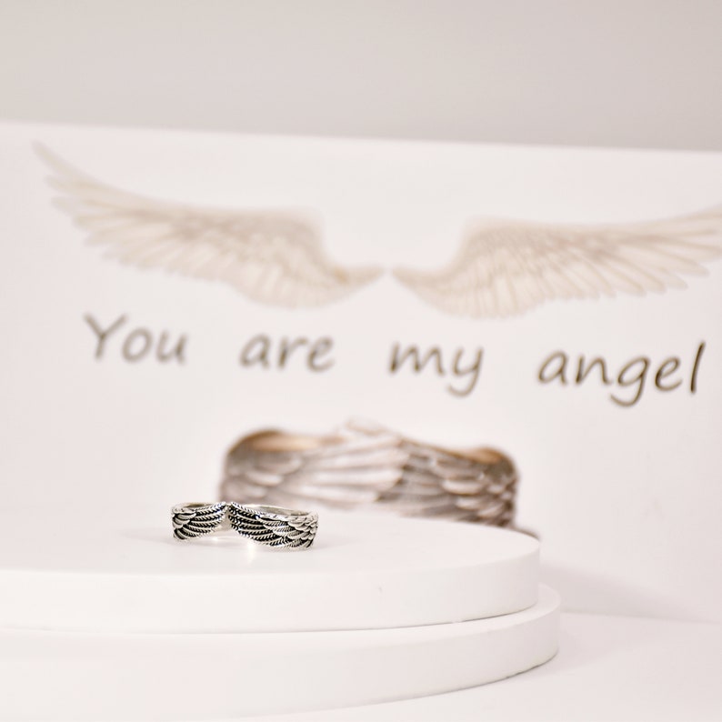 May include: A silver ring with a design of two angel wings. The ring is on a white platform with a card behind it that says "You are my angel".