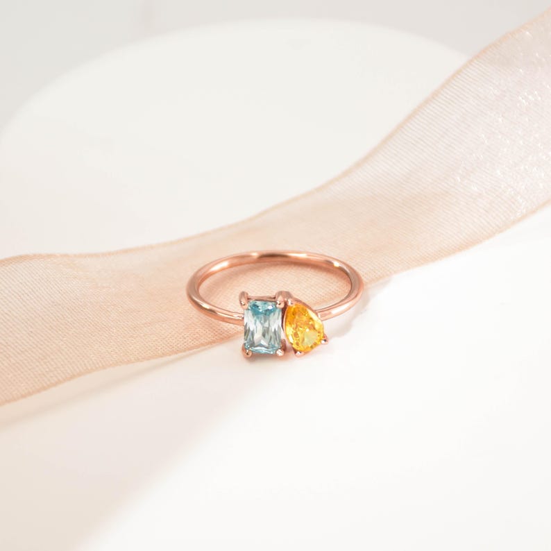 May include: A rose gold ring with a light blue square-shaped gemstone and a yellow pear-shaped gemstone.