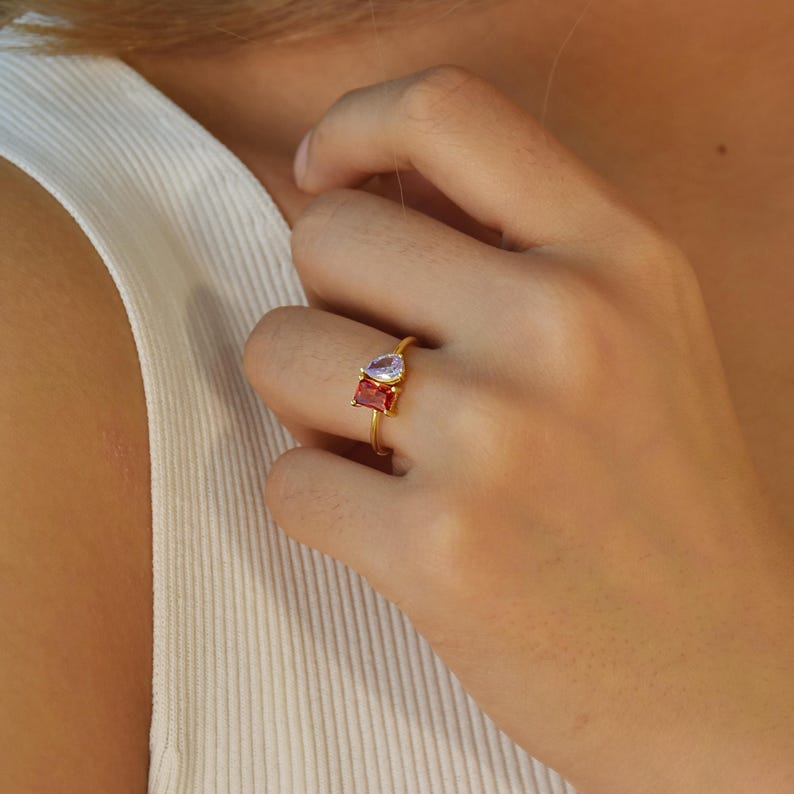 Pu&ograve; includere: Un anello d'oro con una gemma a forma di pera color lavanda e una gemma rossa rettangolare. L'anello &egrave; indossato su un dito, mettendo in risalto il design e il contrasto cromatico del gioiello. La fascia dell'anello &egrave; sottile e delicata.
