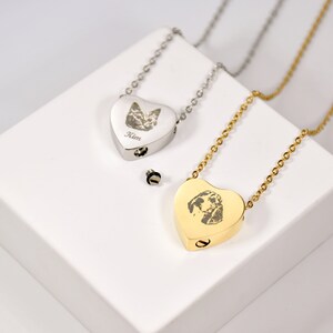 May include: Two heart-shaped pet memorial necklaces. One is silver with a cat portrait and the name "Kim". The other is gold with a dog portrait. Both have a small screw-top compartment and are on delicate chains.