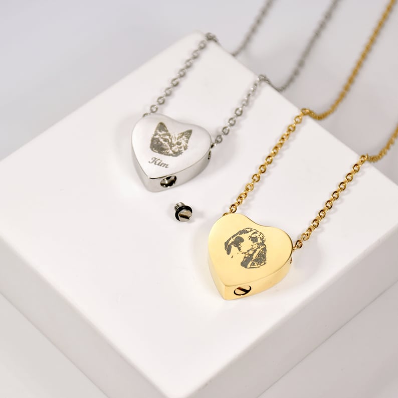 May include: Two heart-shaped pet memorial necklaces. One is silver with a cat portrait and the name "Kim". The other is gold with a dog portrait. Both have a small screw-top compartment and are on delicate chains.