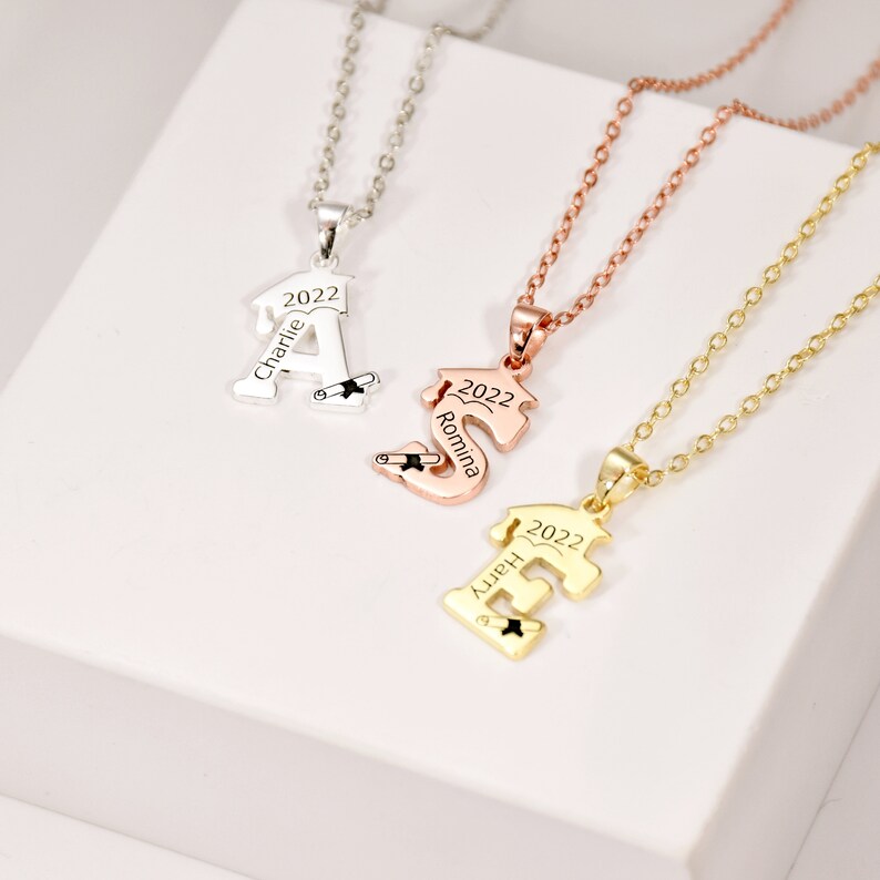 May include: Three gold necklaces with personalized graduation pendants. The pendants are shaped like the letters A, S, and E, and each has a graduation cap and a scroll. The necklaces are on a white surface.