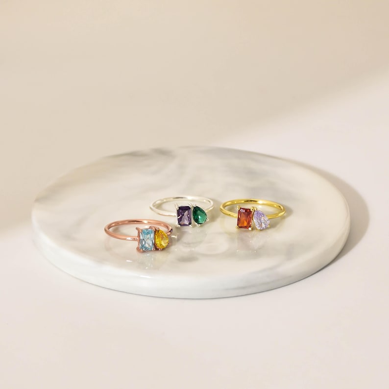 Pu&ograve; includere: Tre anelli con pietre preziose colorate, in oro rosa, argento e oro. L'anello in oro rosa ha pietre blu e gialle. L'anello in argento ha pietre viola e verdi. L'anello in oro ha pietre arancioni e viola. Gli anelli sono su una superficie di marmo.