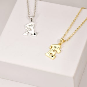 May include: Two graduation necklaces, one silver and one gold, with the letters A and E, the year 2022, and the names Charlie and Happy engraved on them.