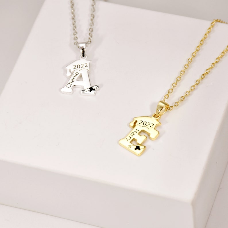 May include: Two graduation necklaces, one silver and one gold, with the letters A and E, the year 2022, and the names Charlie and Happy engraved on them.