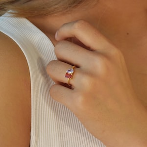 May include: A gold ring with two gemstones. One gemstone is a square, red stone, and the other is a teardrop-shaped, purple stone.