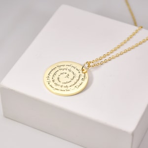 Puede incluir: Un collar con colgante dorado con un colgante circular grabado con una cita en espiral. La cadena es fina y dorada. El collar se muestra sobre una superficie blanca.