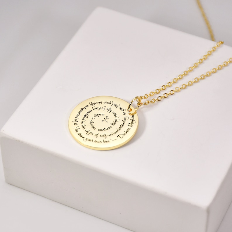 Puede incluir: Un collar con colgante dorado con un colgante circular grabado con una cita en espiral. La cadena es fina y dorada. El collar se muestra sobre una superficie blanca.