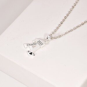 May include: Silver graduation necklace with a charm shaped like a person wearing a graduation cap. The charm is engraved with the name "Charlie" and the year "2022".