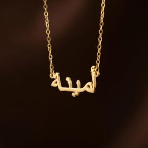 May include: A gold necklace with a fine chain. The pendant displays Arabic script. The necklace is set against a dark brown backdrop, accentuating the gold's gleam and the pendant's design. A piece of jewellery.