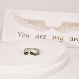 May include: A silver ring with a wing design. The ring is on a white surface with a card that says "You are my angel" in the background.