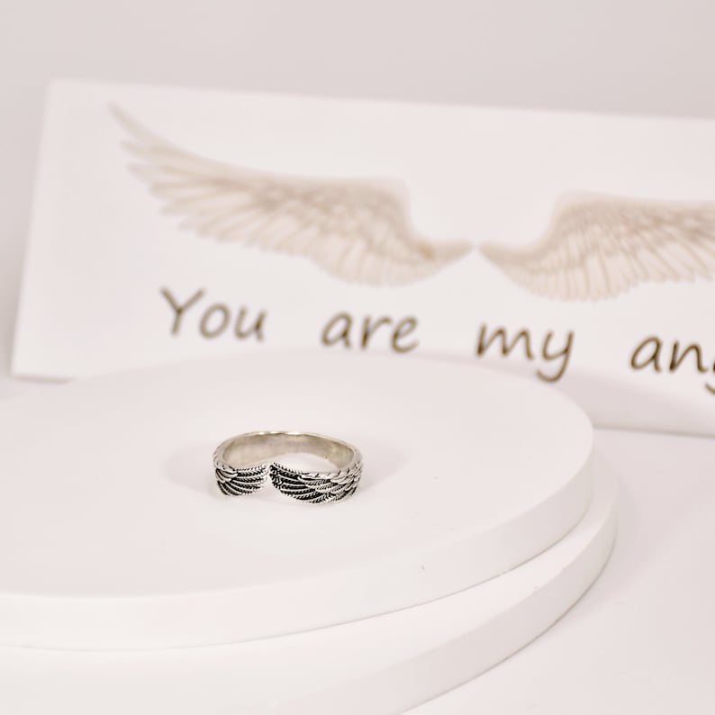 May include: A silver ring with a wing design. The ring is on a white surface with a card that says "You are my angel" in the background.