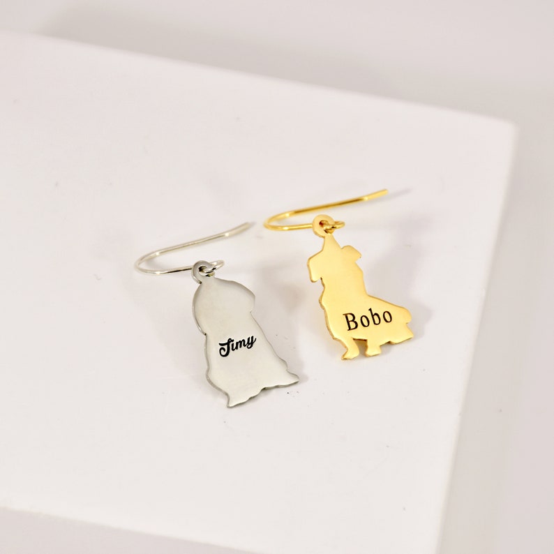 K&ouml;nnte beinhalten: Zwei goldfarbene Ohrh&auml;nger mit einer Hundesilhouette. Die Ohrringe sind mit den Namen "Tiny" und "Bobo" personalisiert.