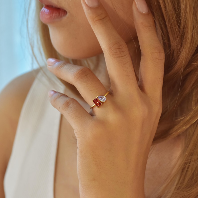 May include: A gold ring with a red and clear gemstone setting. The ring is on a person's finger.