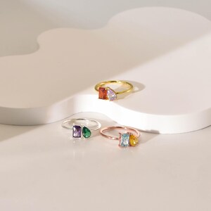 May include: Four rings with different colored gemstones. The rings are made of silver and gold. The gemstones are a light purple, a dark green, a light blue, and a yellow.