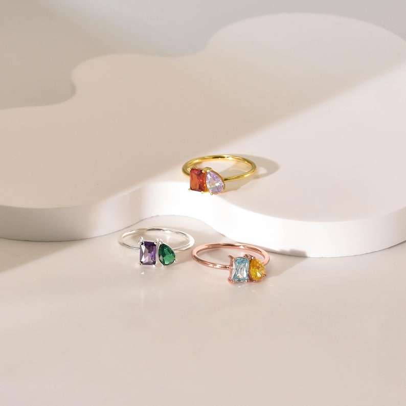 May include: Four rings with different colored gemstones. The rings are made of silver and gold. The gemstones are a light purple, a dark green, a light blue, and a yellow.