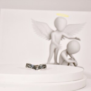 May include: A silver ring with a detailed wing design. The ring is on a white pedestal.