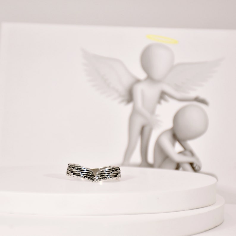 May include: A silver ring with a detailed wing design. The ring is on a white pedestal.