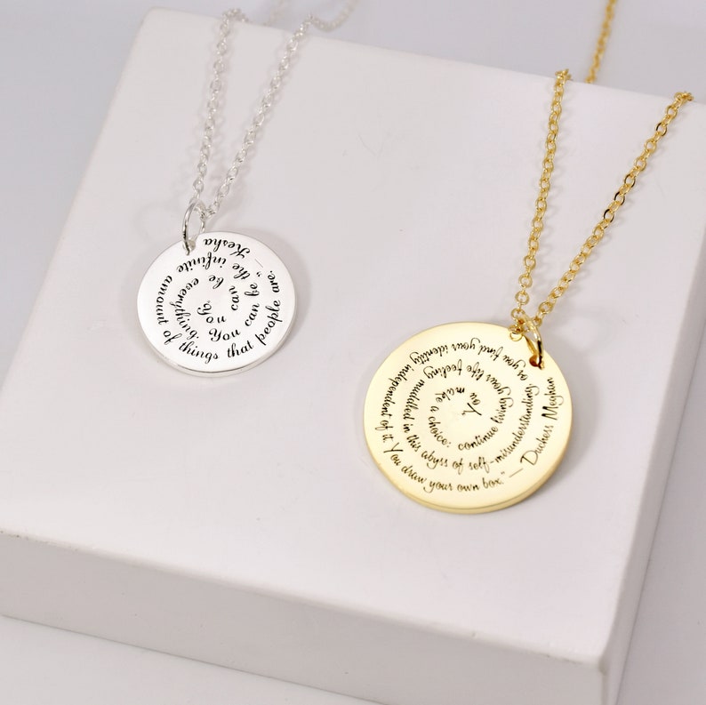 Puede incluir: Dos collares con colgantes circulares. Uno es plateado, el otro dorado. Ambos tienen una cadena y un colgante circular con texto grabado. El colgante plateado dice "Kesha...la infinita cantidad de cosas que la gente puede ser..." El colgante dorado dice "Duquesa Meghan...Encuentras tu identidad..."
