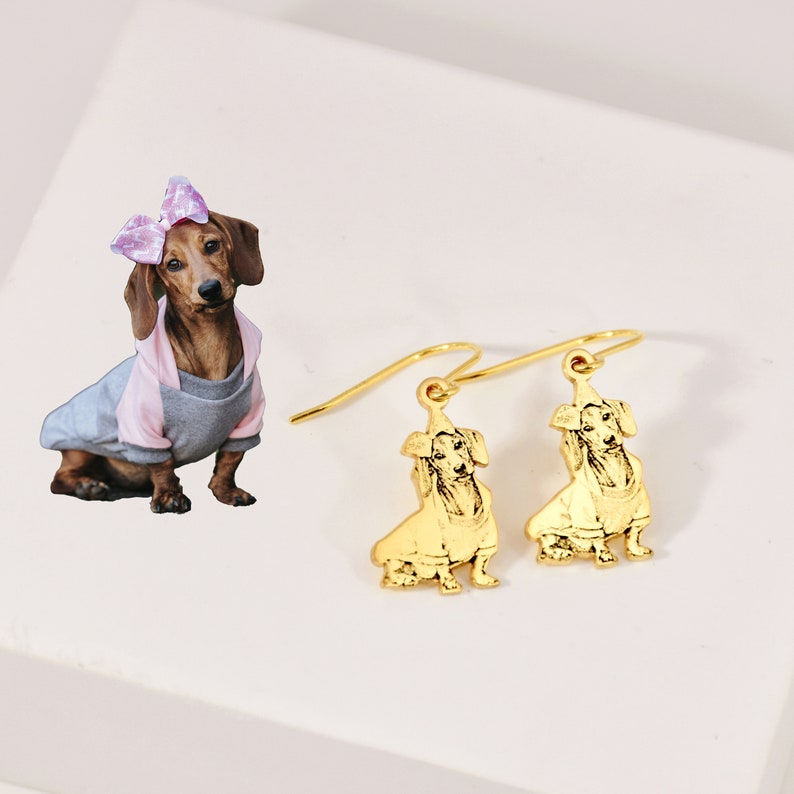 K&ouml;nnte beinhalten: Goldene Ohrh&auml;nger mit einem Dachshund-Silhouetten-Anh&auml;nger. Der Hund tr&auml;gt einen Pullover und eine Schleife auf dem Kopf.