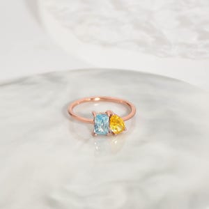 Pu&ograve; includere: Un anello in oro rosa con una gemma rettangolare azzurra e una gemma gialla a forma di pera. L'anello &egrave; appoggiato su una superficie di marmo bianco.