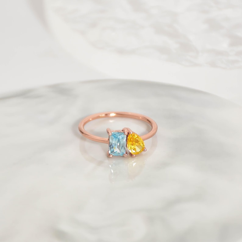 Pu&ograve; includere: Un anello in oro rosa con una gemma rettangolare azzurra e una gemma gialla a forma di pera. L'anello &egrave; appoggiato su una superficie di marmo bianco.