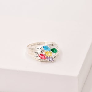 May include: A silver ring with a cluster of colorful gemstones in blue, green, yellow, red, and purple. The ring has the names "Miles", "Tripp", "Alex", "Nora", and "Xander" engraved on it. The words "Nanny's babies" are also engraved on the ring.
