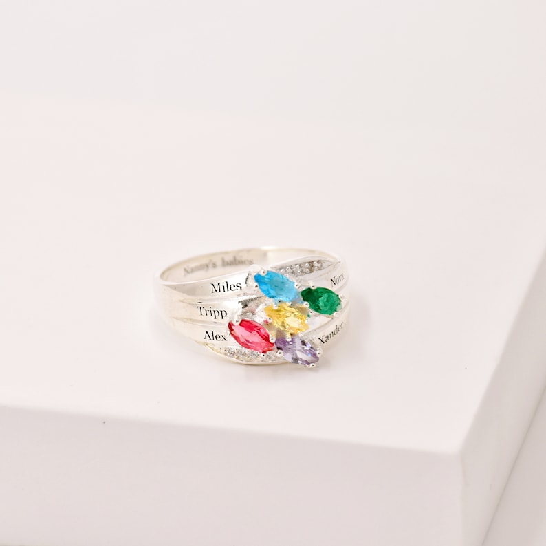 May include: A silver ring with a cluster of colorful gemstones in blue, green, yellow, red, and purple. The ring has the names "Miles", "Tripp", "Alex", "Nora", and "Xander" engraved on it. The words "Nanny's babies" are also engraved on the ring.