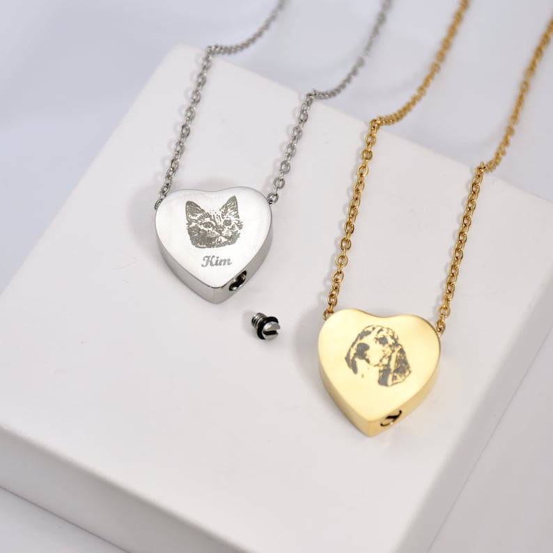 May include: Two heart-shaped pet memorial necklaces. One is silver with a cat portrait and the name "Kim" engraved. The other is gold-coloured with a dog portrait. Both have a small screw-top opening for ashes.